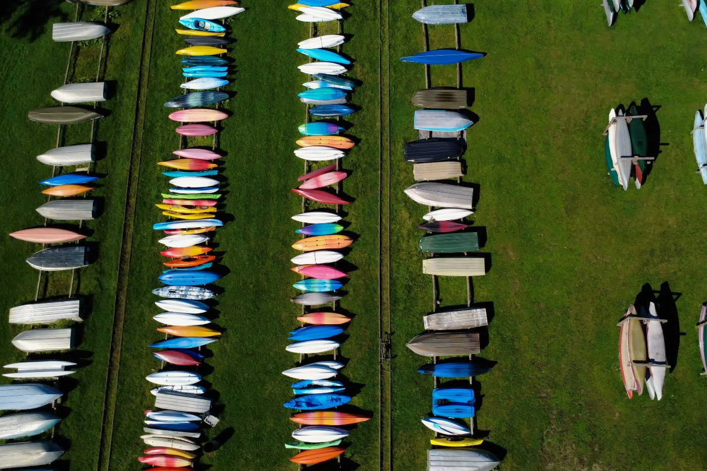 Straight-down aerial view of rows of colorful kayaks and boats on racks across a green lawn