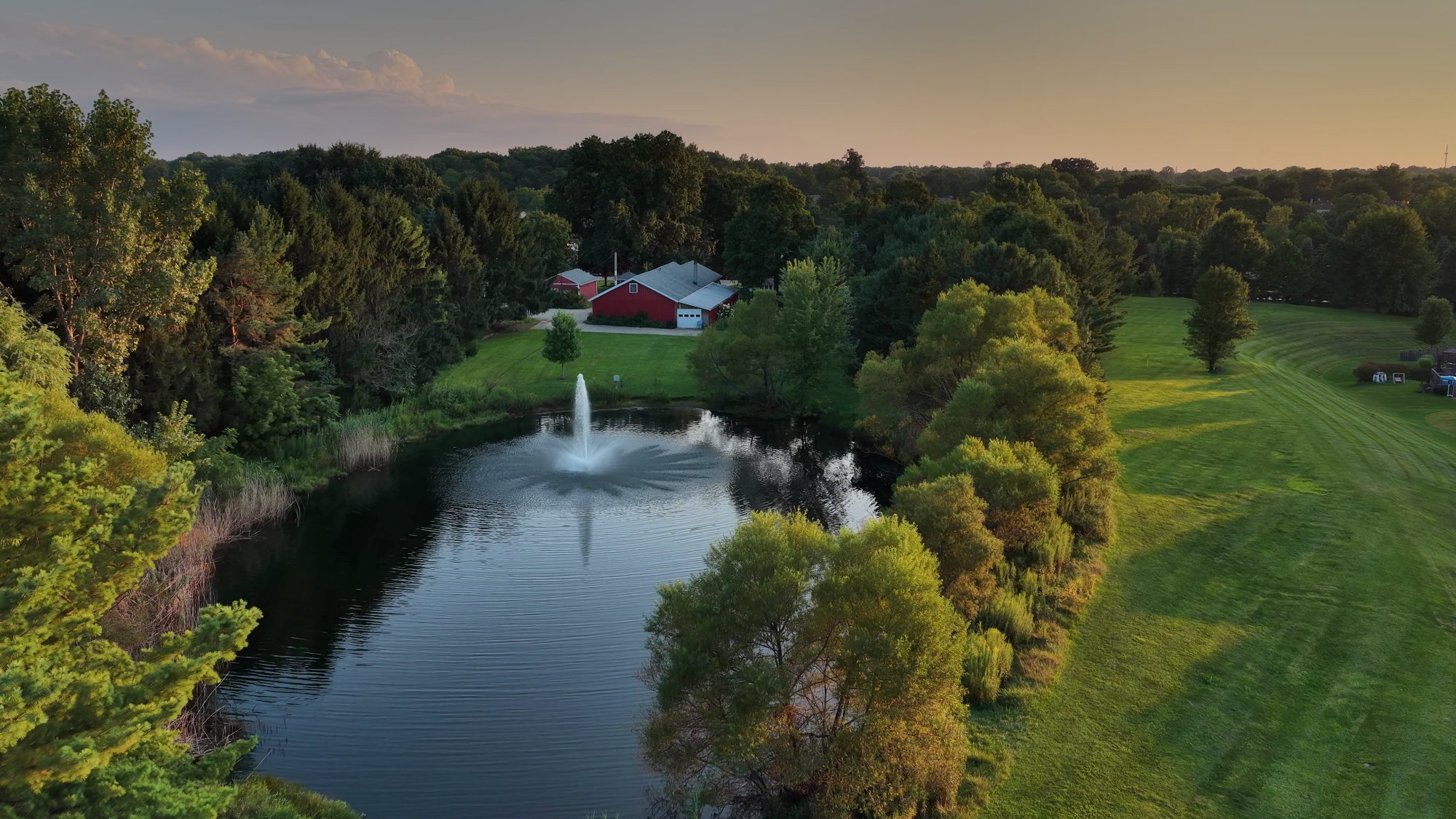 Aerial view at golden hour of a property with a fountain pond, lawn and outbuildings